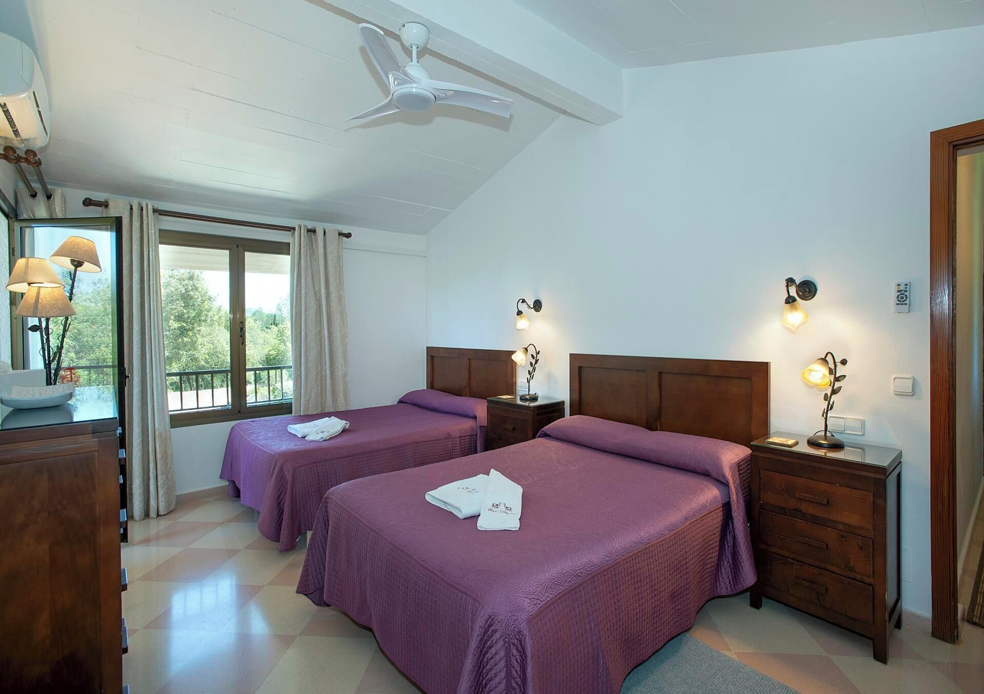 Een hotelkamer met twee bedden, een plafondventilator, een raam met gordijnen, bedlampjes en een houten dressoir.