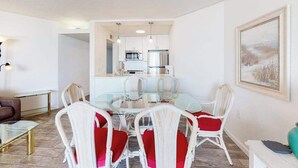 A modern living room with a glass dining table, white wicker chairs with red cushions, a sofa, and a coffee table. - Oceanfront Condo with a Balcony, Great View, AC, WiFi & Shared Outdoor Pool (Ocean City)