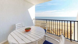 A white table with a bowl on it, a white chair, and a view of the beach through a balcony railing. - Oceanfront Condo with a Balcony, Great View, AC, WiFi & Shared Outdoor Pool (Ocean City)