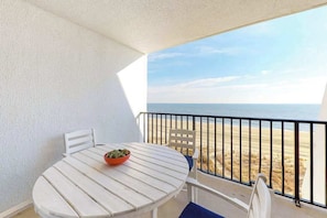 A white table with a bowl on it, a white chair, and a view of the beach through a balcony railing. - Oceanfront Condo with a Balcony, Great View, AC, WiFi & Shared Outdoor Pool (Ocean City)