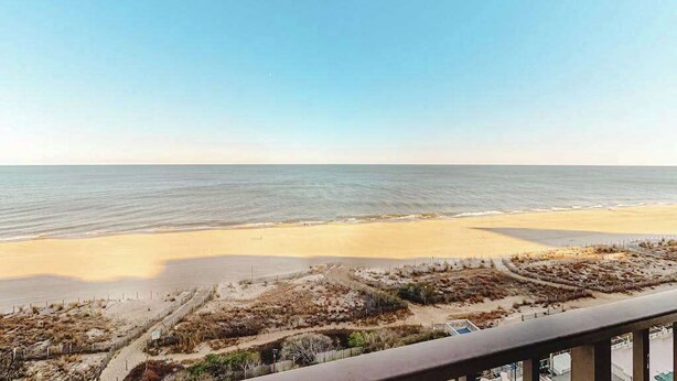 Blick auf einen Strand vom Balkon aus, mit blauem Himmel, Sandstrand und vereinzelter Vegetation.