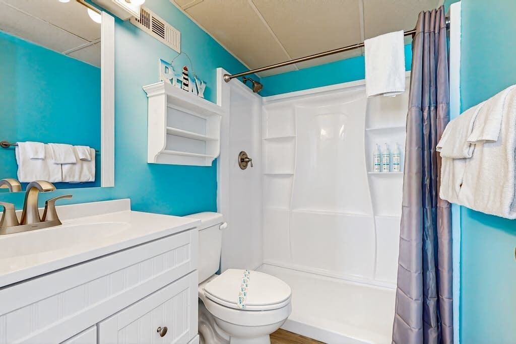 Une salle de bain avec un lavabo, des toilettes et une douche, le tout de couleur blanche. Des serviettes blanches sont accrochées au mur, et il y a un rideau de douche.