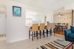 A modern kitchen with white cabinets, a bar area with bar stools, a dining table with chairs, and a blue sofa in a living area. - Ocean Block Condo with Balcony, Partial Ocean View, Central AC & Washer/Dryer (Ocean City)