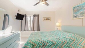 A bedroom with a bed, a ceiling fan, a TV mounted on the wall, and a window with curtains. - Ocean Block Condo with Balcony, Partial Ocean View, Central AC & Washer/Dryer (Ocean City)