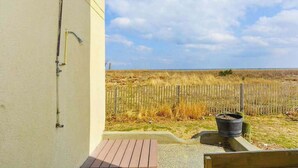 A building with a wooden deck, a metal pipe on the wall, and a view of a grassy field with a wooden fence. - Ocean Block Condo with Balcony, Partial Ocean View, Central AC & Washer/Dryer (Ocean City)
