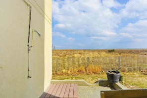 A building with a wooden deck, a metal pipe on the wall, and a view of a grassy field with a wooden fence. - Ocean Block Condo with Balcony, Partial Ocean View, Central AC & Washer/Dryer (Ocean City)