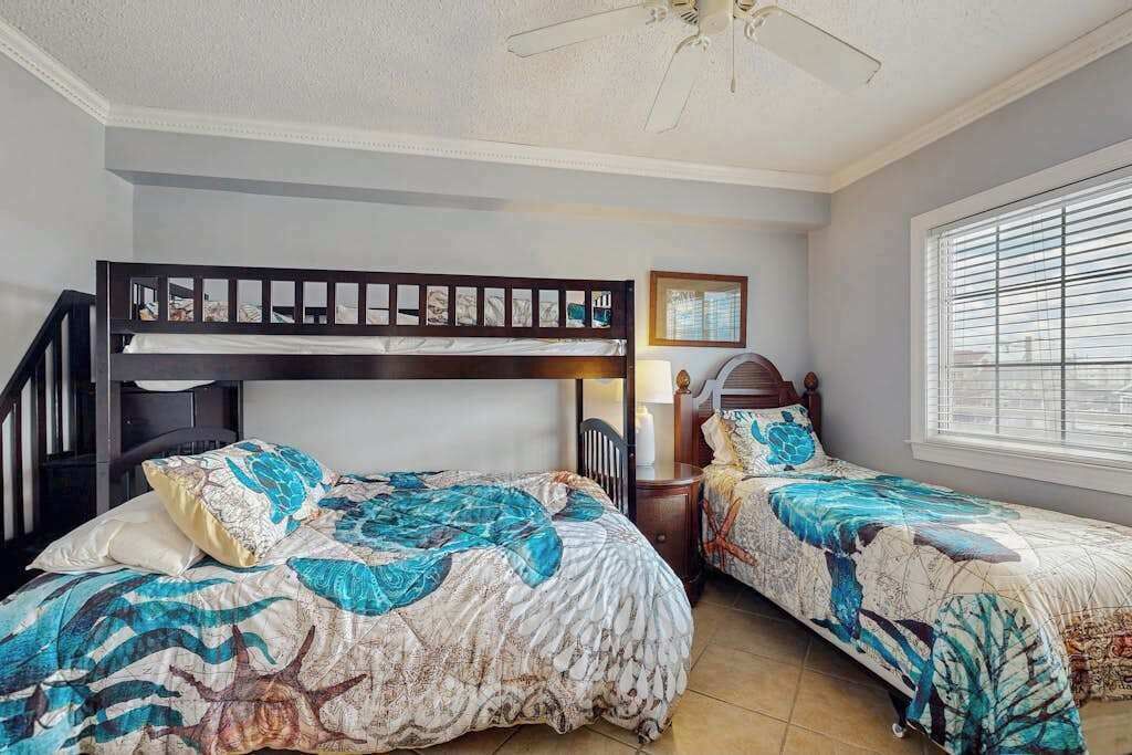 A bedroom with bunk beds, a ceiling fan, a window with blinds, and a bedside table with a lamp.