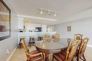 A dining area with a round table and six chairs, a kitchen in the background, and a framed picture on the wall. - Lovely beachfront condo with ocean view, pool, balcony, fireplace, AC, & W/D (Ocean City)