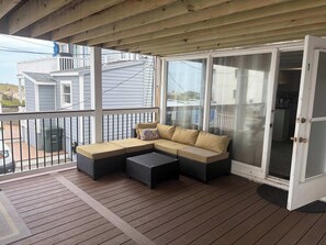 A covered deck with a beige sectional sofa, a small table, and a sliding glass door leading to a kitchen. - Convenient Condo, Updated Waterfront Deck, Free WiFi, Games, Partial A/C, W/D (Ocean City)