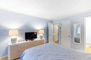 Ein Schlafzimmer mit Bett, Fernseher, Kommode und einer Tür zu einem weiteren Raum.