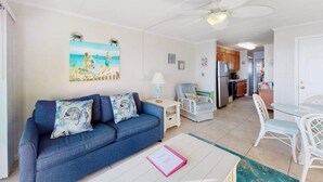 A living room with a sofa, a dining table with chairs, a ceiling fan, and a kitchen area in the background. - Air-Conditioned Condo Steps from the Beach w/ Balcony, WiFi, Washer/Dryer (Ocean City)