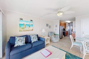 Ein Wohnzimmer mit einer Couch, einem Esstisch mit Stühlen, einem Deckenventilator und einer Küchenzeile im Hintergrund.