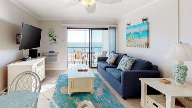 A room with a TV, a sofa, a coffee table, a ceiling fan, and a view of the ocean. - Air-Conditioned Condo Steps from the Beach w/ Balcony, WiFi, Washer/Dryer (Ocean City)