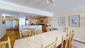 A spacious dining area with a marble table and wooden chairs, a kitchen with wooden cabinets, and framed pictures on the wall. - Incredible Beachfront Condo w/Private Balcony, Fireplace, A/C, & Shared Pool (Ocean City)