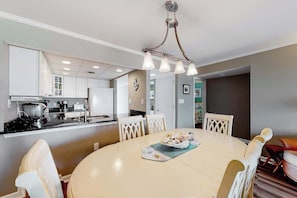 A dining area with a round table, chairs, a chandelier, and a kitchen in the background. - Beachfront Condo w/Private Balcony, Beach Views, Washer/Dryer, Central A/C (Ocean City)