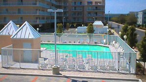 Ein Schwimmbad mit umzäunter Liegefläche, Liegestühlen und einem Gebäude im Hintergrund.
