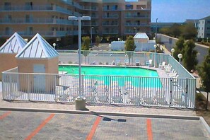 Ein Schwimmbad mit umzäunter Liegefläche, Liegestühlen und einem Gebäude im Hintergrund.