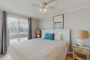 A bedroom with a large bed, a ceiling fan, a nightstand with a lamp, and a window with curtains. - Makai 108 (Ocean City)