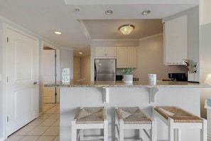 A kitchen with white cabinets, a stainless steel refrigerator, a granite countertop, and three bar stools with woven seats. - Makai 108 (Ocean City)