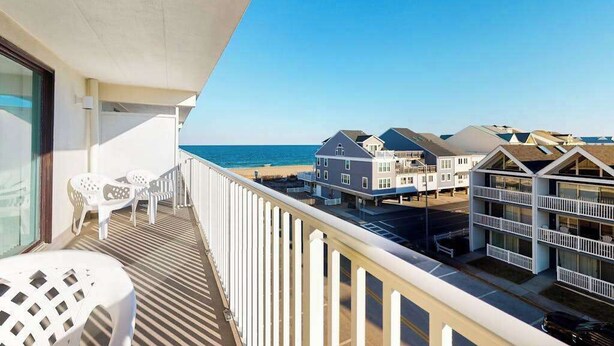 A balcony with a view of the beach and ocean, white plastic chairs, and a building with multiple balconies. - Ocean Block Condo w/ Side Ocean View, Balcony, WiFi, AC & Shared Pool (Ocean City)