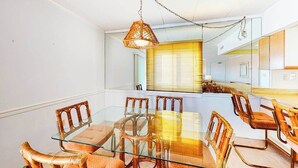 A dining area with a glass table and wicker chairs, a hanging light fixture, and a kitchen in the background. - Ocean Block Condo w/ Side Ocean View, Balcony, WiFi, AC & Shared Pool (Ocean City)