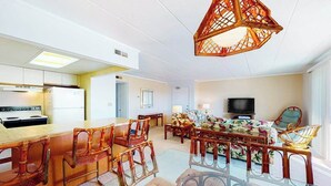 A modern kitchen with a refrigerator, a dining area with a glass table and wooden chairs, and a living room with a sofa and a television. - Ocean Block Condo w/ Side Ocean View, Balcony, WiFi, AC & Shared Pool (Ocean City)