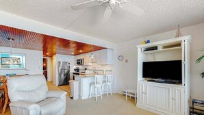 Ein Wohnzimmer mit einem weißen Sofa, einem Fernseher auf einem weißen Schrank, einem Deckenventilator und einer Küche im Hintergrund.