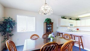 Un coin repas avec une table en verre, des chaises en bois, un lustre, une plante en pot et une vue sur la cuisine.