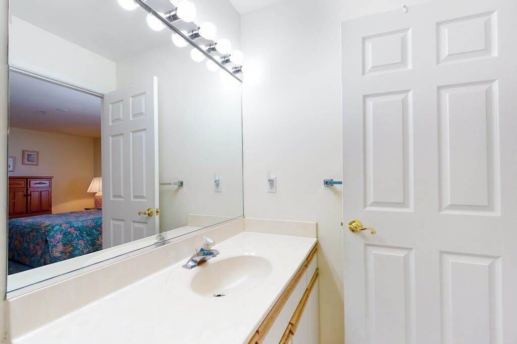A bathroom with a white sink, mirror, and vanity, a door with a brass handle, and a light fixture above the mirror.