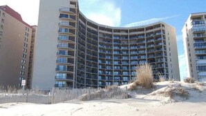 Un hotel de varios pisos con fachada curva, balcones y un área de playa arenosa al frente.