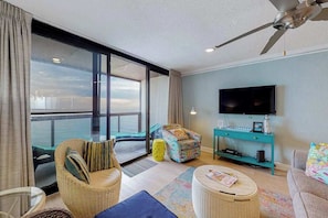 A modern living room with a large window, a ceiling fan, a flat-screen TV, and a teal console table. - Updated waterfront condo with balcony, pools, game room, sauna & gym (Ocean City)