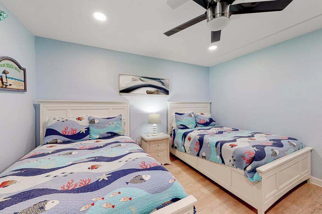 A bedroom with two beds, a ceiling fan, a nightstand with a lamp, and a wall-mounted artwork.