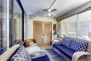 A room with a ceiling fan, a sofa with patterned cushions, a wicker chair, and a large window. - Beachfront condo with amazing views, balcony, & outdoor pool (Ocean City)