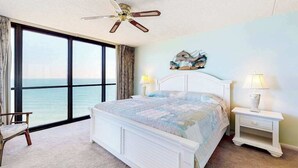 A bedroom with a large window, a bed, a chair, a nightstand, and a ceiling fan. - Budget-friendly beachfront condo with balcony, pools, gym & sauna (Ocean City)