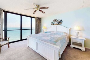 Ein Schlafzimmer mit großem Fenster, einem Bett, einem Sessel, einem Nachttisch und einem Deckenventilator.
