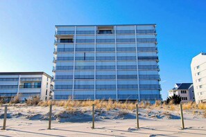 A multi-story building with a grid-like facade, situated on a sandy area with sparse vegetation. - Inviting beachfront condo with beach views, balcony, AC & washer/dryer (Ocean City)