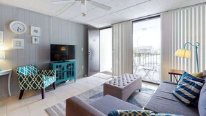 A modern living room with a ceiling fan, a television, a sofa, a chair, and a sliding glass door leading to an outdoor area. - Grandview 203 (Ocean City)