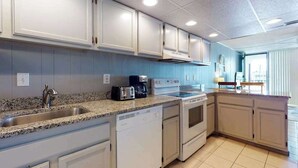 A kitchen with white cabinets, a granite countertop, a dishwasher, a stove, and a coffee maker. - Grandview 203 (Ocean City)