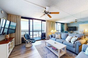 A modern living room with a ceiling fan, a television, a sofa, a coffee table, and a view of the ocean through large windows. - Affordable beach condo with balcony views, pools & game room (Ocean City)
