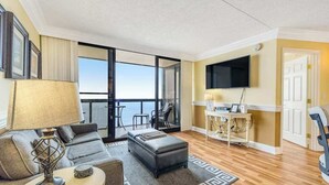 A modern living room with a sofa, ottoman, and a table with a lamp. There is a balcony with a view, a flat-screen TV mounted on the wall, and a door leading to another room. - Cozy beachfront condo with balcony, washer/dryer, pools & gym (Ocean City)