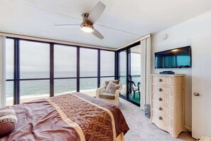 Ein Schlafzimmer mit einem Bett, einem Sessel, einer Kommode, einem Fernseher und einem großen Fenster mit Meerblick.
