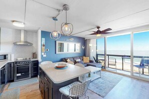 A modern kitchen with a central island, a dining area with a view, and a large window overlooking the beach. - Inviting & modern beachfront condo with balcony, AC, pools, gym & tennis (Ocean City)