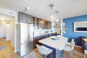 A modern kitchen with stainless steel appliances, a central island with bar stools, and a blue accent wall. - Inviting & modern beachfront condo with balcony, AC, pools, gym & tennis (Ocean City)