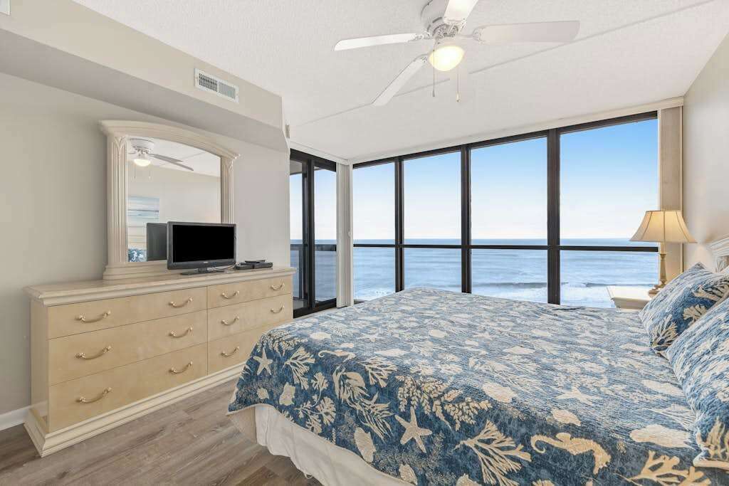 A bedroom with a large bed, a dresser, a television, and a view of the ocean.