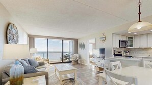 A spacious living area with a sofa, coffee table, and dining table, a kitchen with white cabinets, and a large window offering a view. - Convenient beachfront condo with balcony, pools, tennis, gym & central AC (Ocean City)