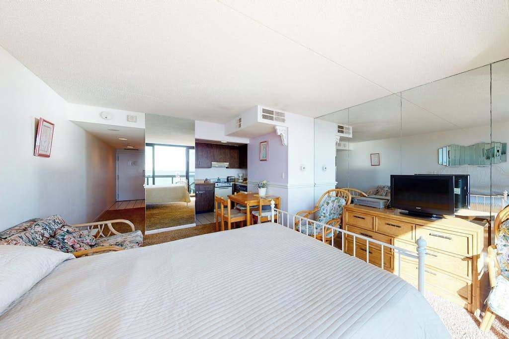 A hotel room with a large bed, a desk with a television, a dining area, and a kitchen in the background.