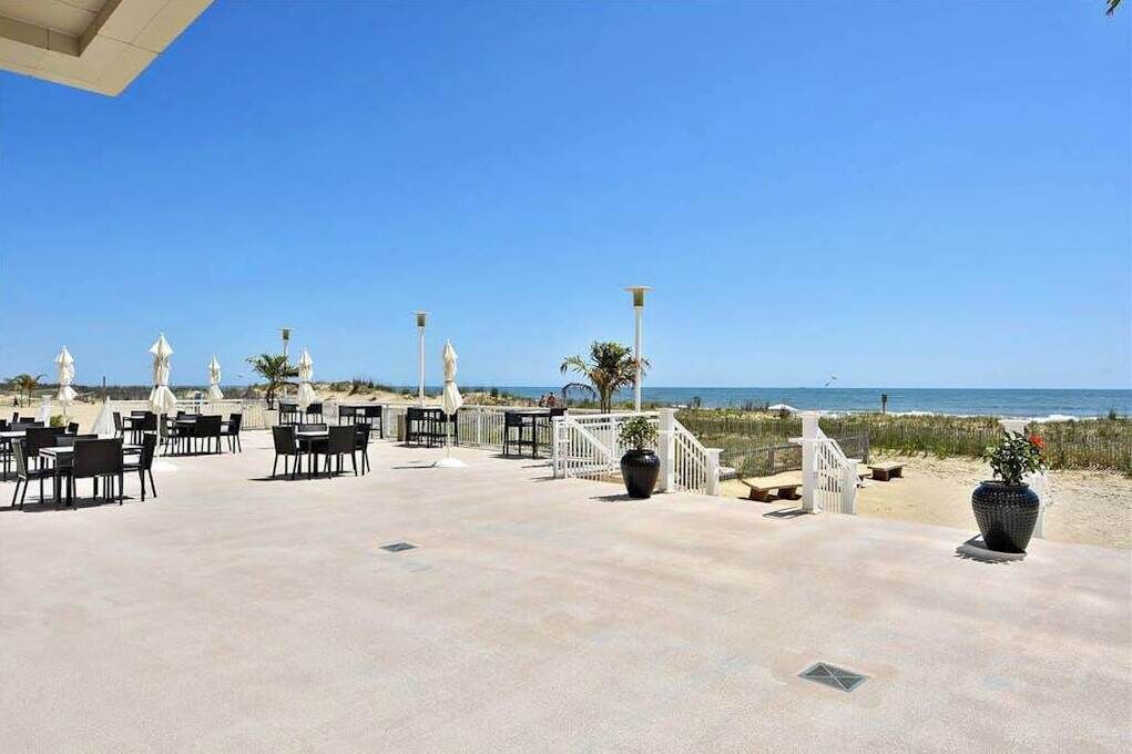 Une terrasse extérieure avec des tables et des chaises, une clôture blanche et une vue sur la plage et l’océan.