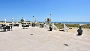 Een terras buiten met tafels en stoelen, een wit hek en uitzicht op het strand en de zee.