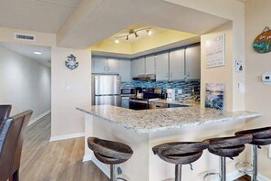 A modern kitchen with a central island, bar stools, and a dining area with chairs. - Adorable beachfront condo with balcony, incredible views, pools & game room (Ocean City)
