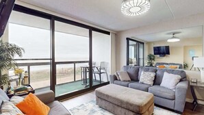 Ein geräumiges Wohnzimmer mit großem Fenster, einem grauen Sofa, einem Hocker und einem Flachbildfernseher.